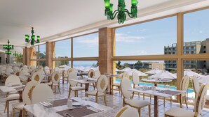 A restaurant with tables and chairs arranged for dining, large windows offering a view of the pool and buildings, and green chandeliers hanging from the ceiling. - Mythos Panorama (Corfu)