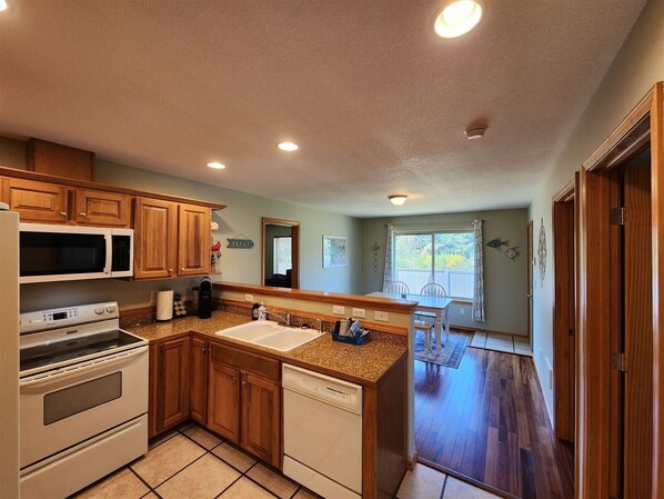A compact kitchen with wooden cabinets, a microwave, an oven, a sink, and a dishwasher. There is a window with curtains and a view of the outdoors.