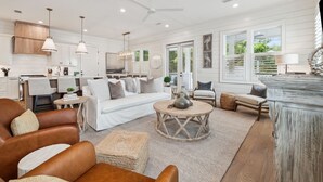 A spacious living room with a white sofa, leather armchairs, and a coffee table. There is a kitchen area with white cabinets and a dining table. The room has large windows with blinds and a ceiling fan. - Private Pool | LSV | Bikes (Santa Rosa Beach)