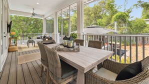 A covered outdoor patio with a wooden table and wicker chairs, large windows offering a view of trees. - Private Pool | LSV | Bikes (Santa Rosa Beach)