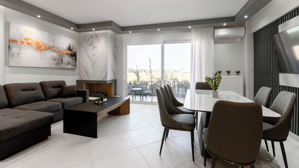 Ein modernes Wohnzimmer mit einer Couch, einem Couchtisch und einem Essbereich mit Stühlen. Ein großes Fenster mit Vorhängen, ein Kamin und ein an der Wand befestigtes Fernsehgerät sind ebenfalls vorhanden.