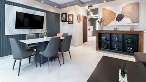 Ein modernes Wohnzimmer mit Flachbildfernseher, einem Essbereich mit weißem Tisch und grauen Stühlen sowie einem dekorativen Regal mit Pflanzen und Figuren.