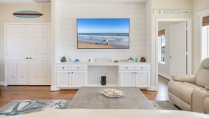 Ein Wohnzimmer mit einem großen Fernseher, der eine Strandansicht zeigt, einem weißen Schrank mit Dekorationen, einem beigen Sofa und einem Couchtisch.