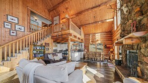 A spacious log cabin interior with a high ceiling, a central staircase, a cozy living area with a sofa and armchairs, and a stone fireplace. - The Broadmeadow Cabin - Vr-25-0005 3 Bedroom Cabin (Lyons)
