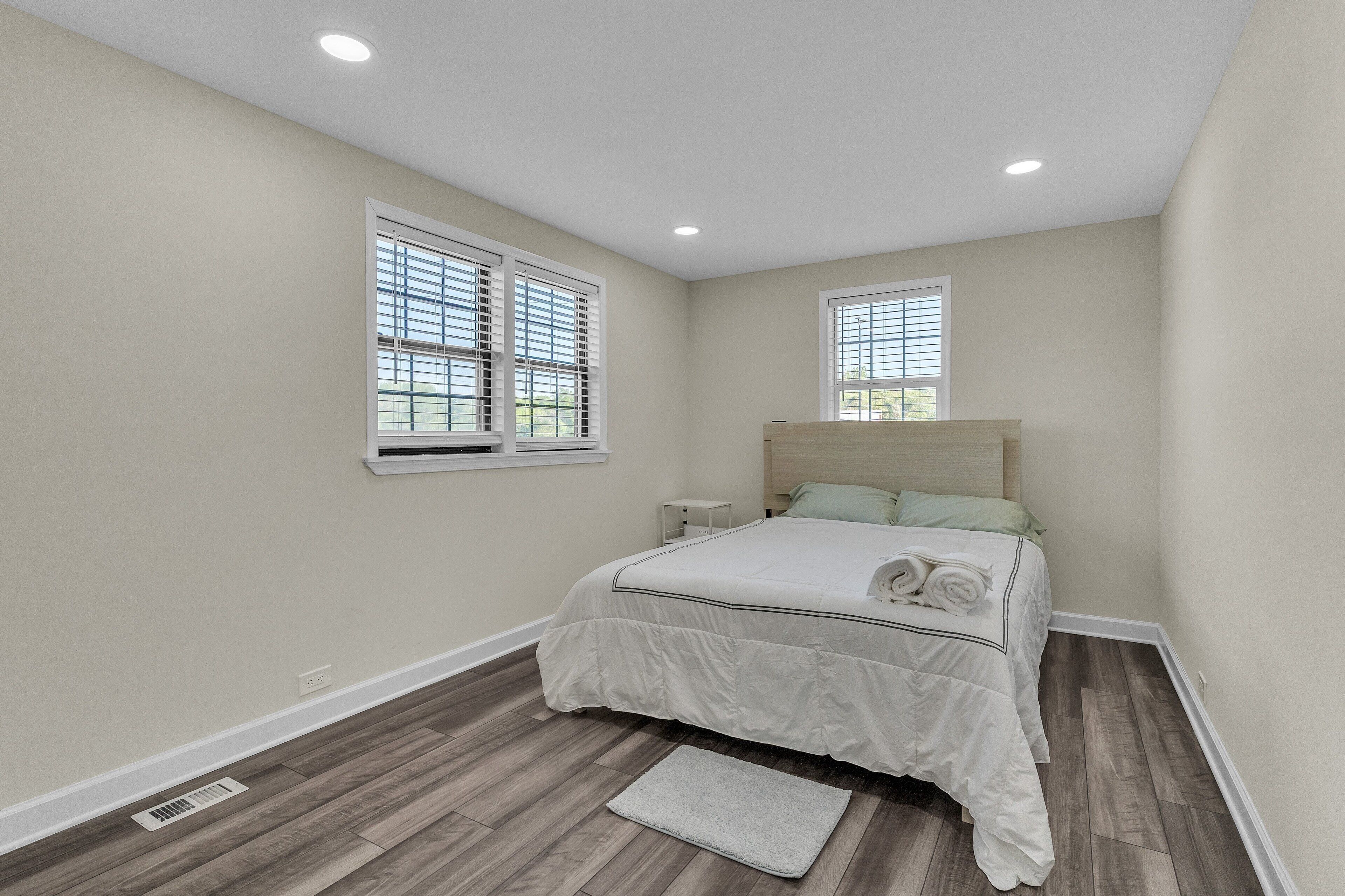 Un dormitorio con una cama, dos ventanas, una mesita de noche y una lámpara.