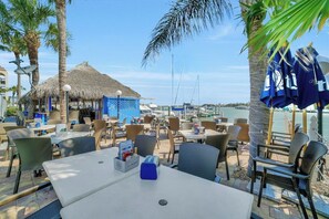 An outdoor dining area with tables and chairs, a thatched-roof structure, and a view of the sea.