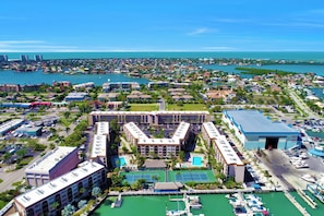 An aerial view of a coastal city with a prominent hotel featuring a central courtyard, tennis courts, and a marina with boats.