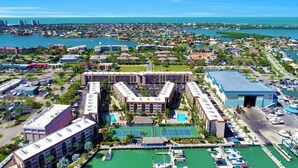 Vista aérea de una ciudad costera con un hotel destacado que cuenta con un patio central, canchas de tenis y un puerto deportivo con barcos.