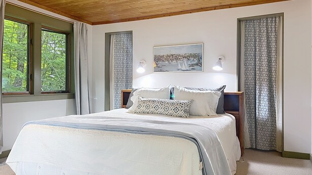 A bedroom with a wooden ceiling, a large bed with patterned pillows, a nightstand, a wall-mounted lamp, and a window with curtains. - Mountainside Getaway (Burke)