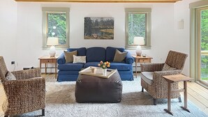 A cozy living room with a blue sofa, a wicker chair, a small table, and two lamps. There are two windows with views of greenery. - Mountainside Getaway (Burke)