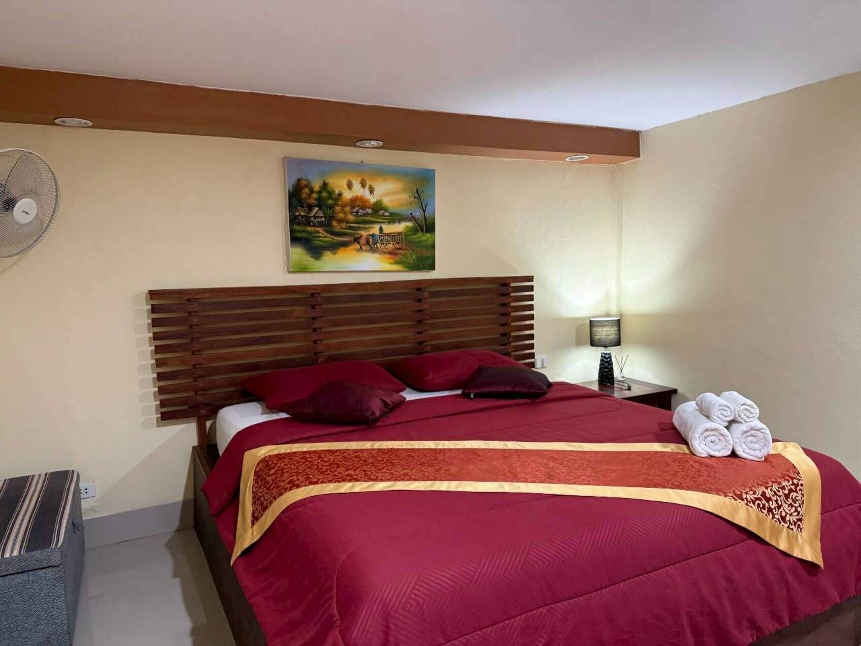 A hotel room with a large bed, a wooden headboard, a painting on the wall, a bedside table with a lamp, and a small table with towels.
