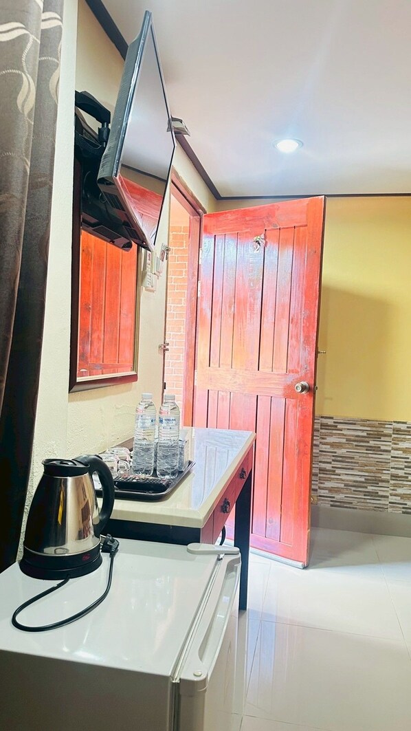 A compact kitchen with a white countertop, a kettle, and a water dispenser. A red door and a window with a curtain are visible in the background.