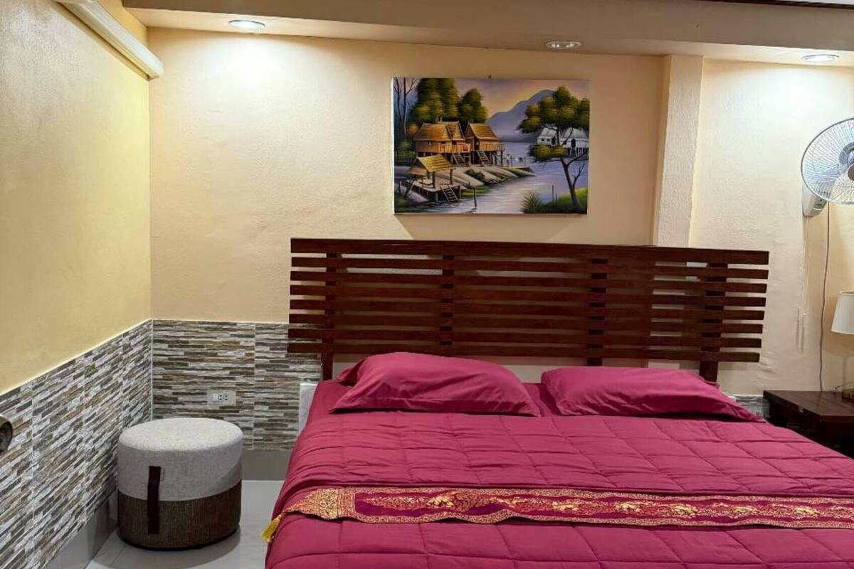 A bedroom with a bed, a painting, a wooden headboard, a round ottoman, and a nightstand with a lamp.