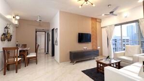 A modern living room with a dining area, a flat-screen TV mounted on the wall, a ceiling fan, and a large window with curtains. - 2BHK Master Suite (Gagal Home) (Mumbai)