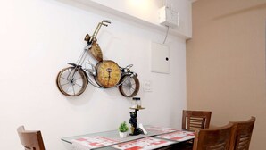 A dining area with a wooden table and four chairs, a wall-mounted bicycle clock, and a wall-mounted light fixture. - 2BHK Master Suite (Gagal Home) (Mumbai)