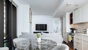 A modern living room with a round marble table, grey upholstered chairs, a grey sofa, a flat-screen TV, and a kitchen with white cabinets and a marble backsplash.