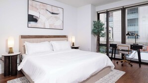 A modern hotel room with a large bed, bedside lamps, a desk with a chair, and a large window offering a city view.