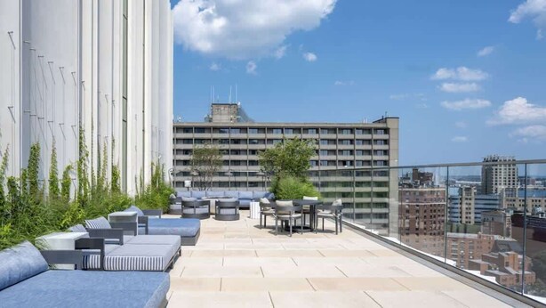 Una terraza en la azotea con mobiliario exterior, vistas de un paisaje urbano y un edificio moderno al fondo.