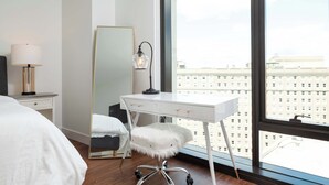 Una habitación de hotel moderna con un ventanal grande, un escritorio blanco con espejo, un sillón mullido y una mesita de noche con lámpara.