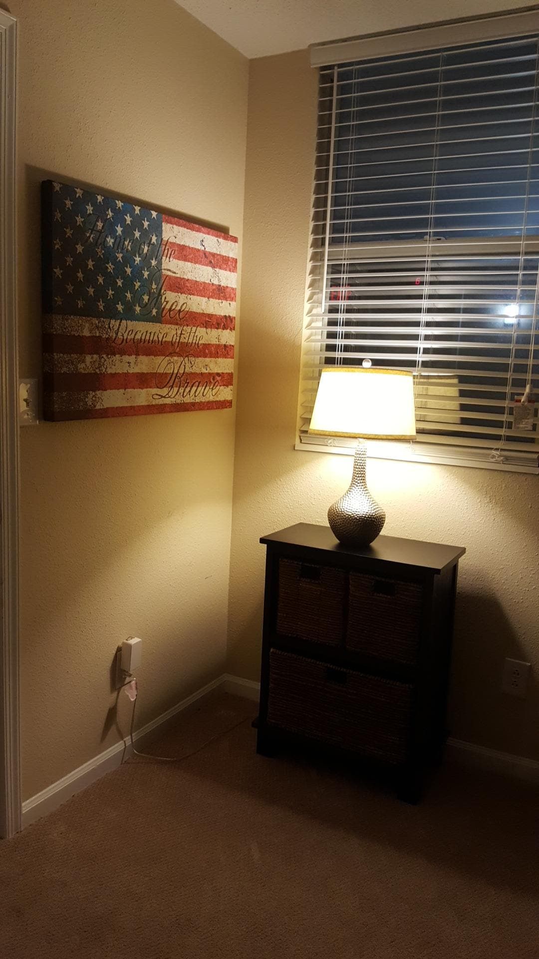 Quarto com bandeira americana fixada na parede, janela com persianas, luminária sobre um armário de madeira e piso coberto de carpete.