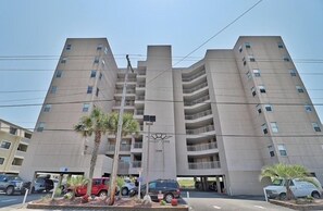 A multi-story building with a central entrance, balconies, and a parking area with vehicles. - Oceanfront 3BR Family Condo with Pool and Views (North Myrtle Beach)