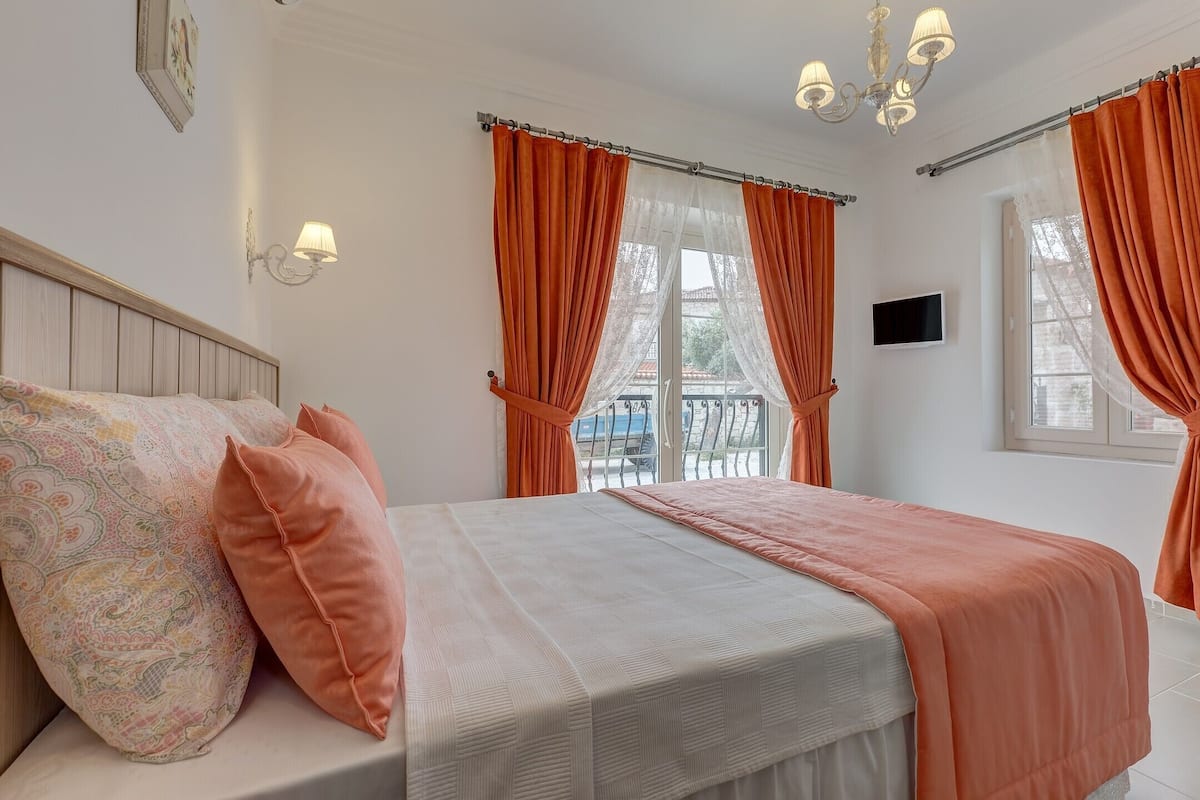 A neatly made bed with a patterned pillow and a solid orange pillow, a white headboard, and a chandelier.