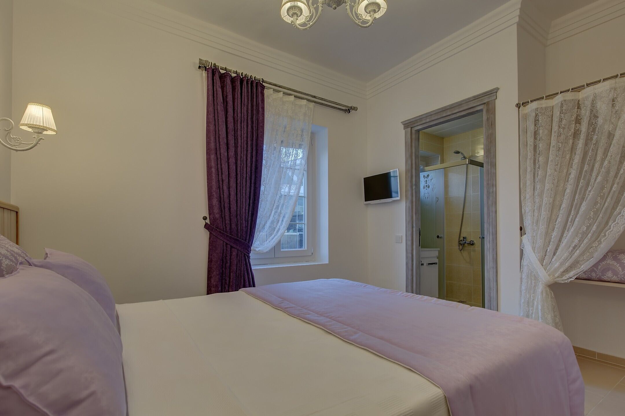 A bedroom with a bed, a window with purple curtains, a shower area, and a TV on the wall.