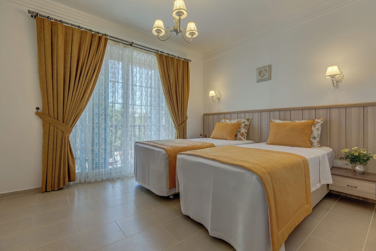 A hotel room with two beds, beige curtains, a chandelier, and a nightstand with a vase of flowers.