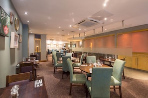A hotel dining area with tables and chairs, a kitchen area, and a reception desk. - Premier Inn Liverpool City Centre - Moorfields (Liverpool)