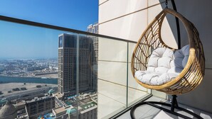 Un balcón alto con una silla de mimbre, barandilla de cristal y vista a una ciudad y al agua.
