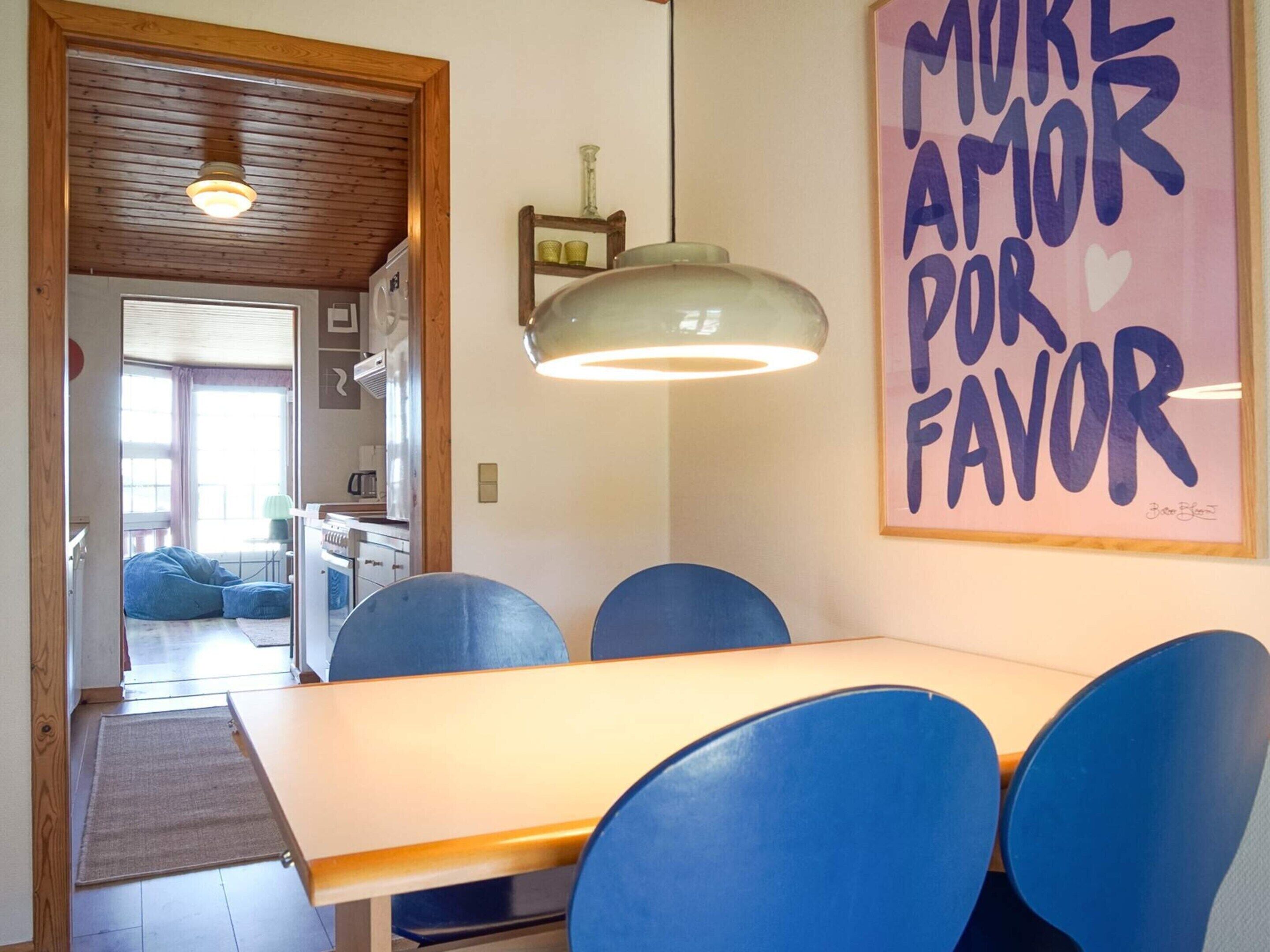 Comedor con mesa y sillas azules, cocina al fondo y una pared con un letrero que dice 'More Amor por Favor'.