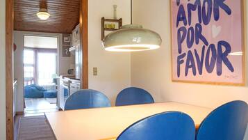 Comedor con mesa y sillas azules, cocina al fondo y una pared con un letrero que dice 'More Amor por Favor'.