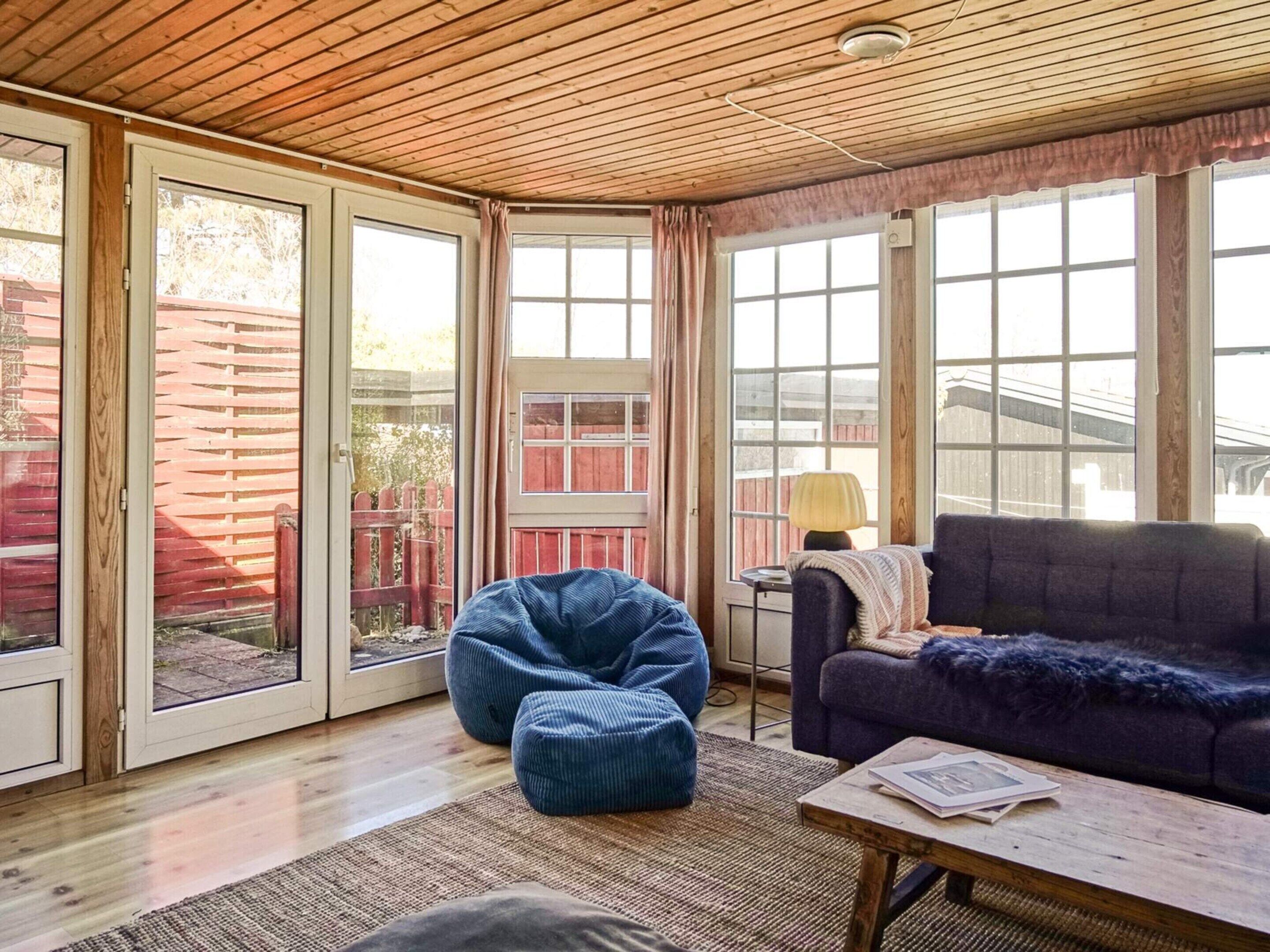 A room with a wooden ceiling, large windows, a blue bean bag, a purple sofa, and a small table with a lamp.