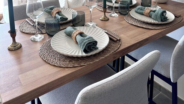 Mesa de comedor dispuesta con platos, vasos y una vela, rodeada de sillas y un sofá en una habitación luminosa con una planta y obras de arte en la pared.