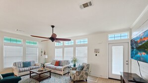 Un ampli saló amb ventilador de sostre, grans finestres amb persianes, televisió i terra de fusta.