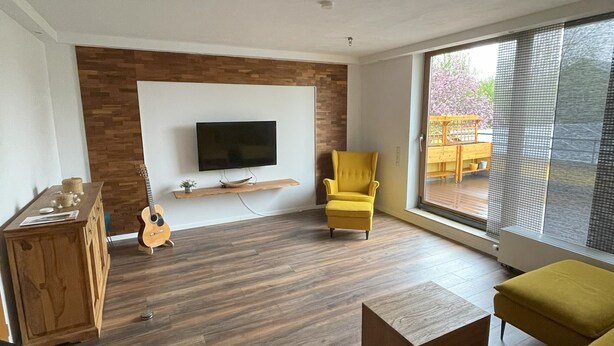 A modern living room with a flat-screen TV mounted on a wooden wall, a yellow armchair, a wooden coffee table, and a yellow sofa. - Vacation home in Eschweiler - relaxation near the Eifel (Eschweiler)