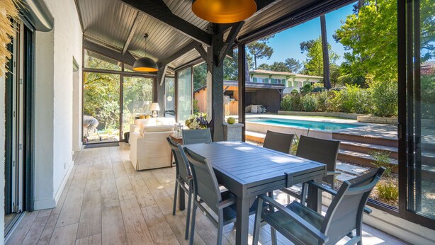 A modern outdoor dining area with a pool view, featuring a wooden deck, metal-framed chairs, and a glass door leading to a pool. - Villa familiale climatisée avec Piscine (Pyla-sur-Mer)