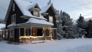 A two-story house with a snow-covered roof and front porch, featuring a Christmas wreath and lights. - In Town Delight! (Manchester)