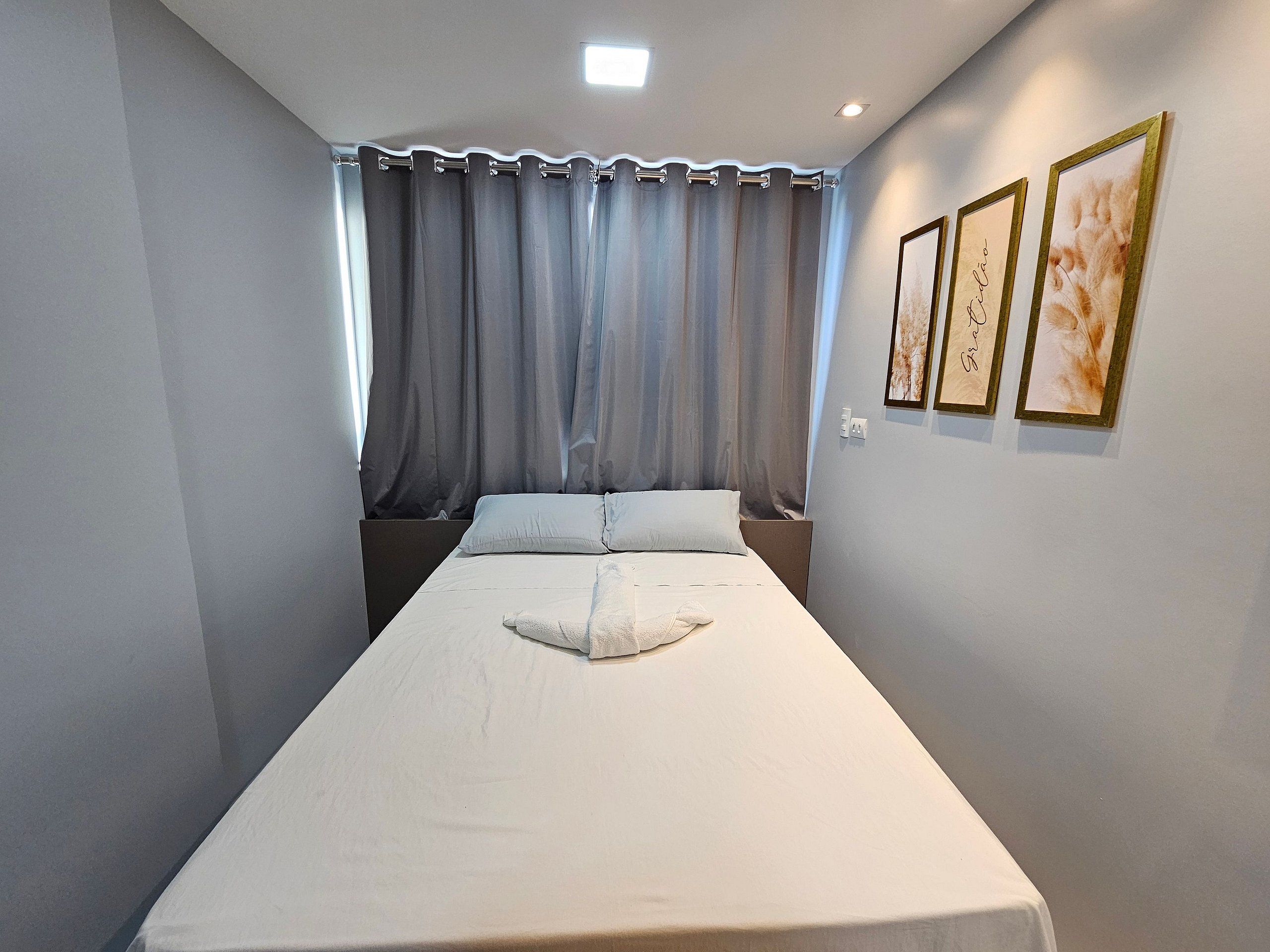 A single bed with a white mattress and pillows, a window with grey curtains, and two framed pictures on the wall.