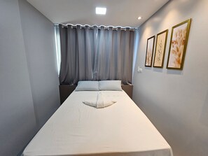 A single bed with a white mattress and pillows, a window with grey curtains, and two framed pictures on the wall. - Eco Mirage Residence (Ipojuca)