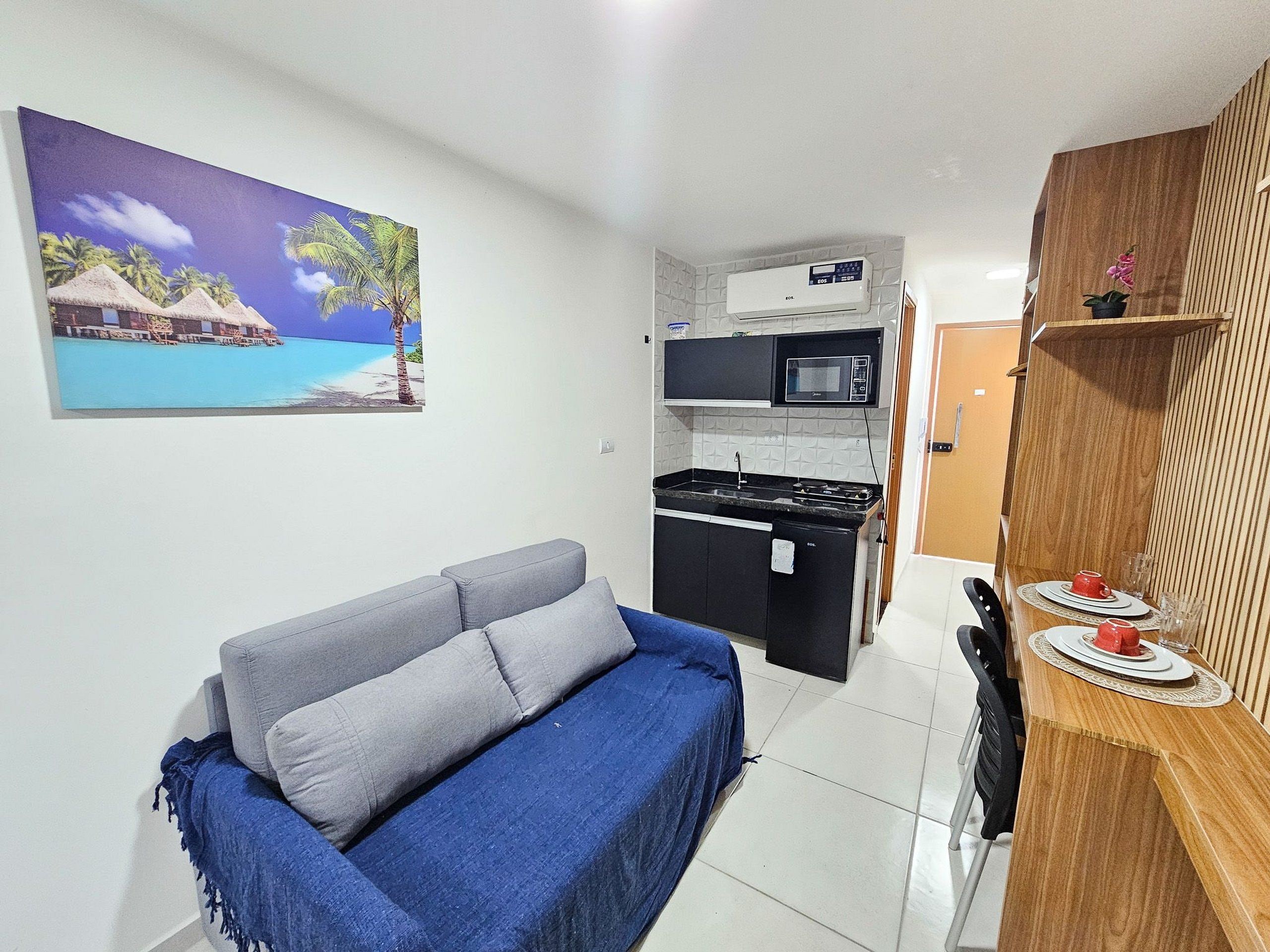 A compact living space with a kitchenette, a sofa, a dining table with red cups, and a painting of a tropical beach.