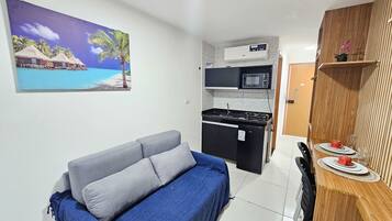 A compact living space with a kitchenette, a sofa, a dining table with red cups, and a painting of a tropical beach.