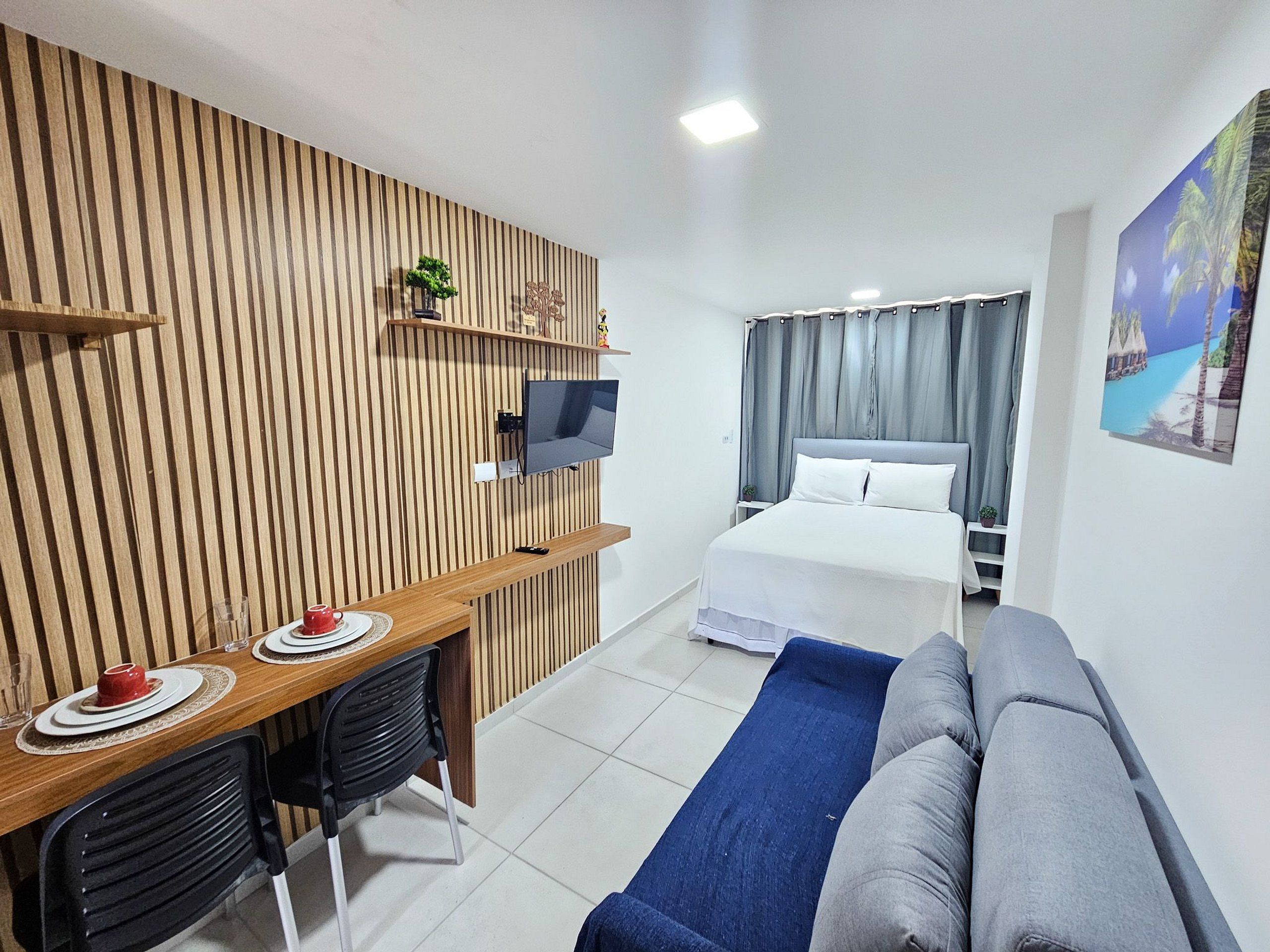A hotel room with a wooden slat wall, a TV mounted on it, a bed with white linens, a blue sofa, and a small table with a red cup and saucer.