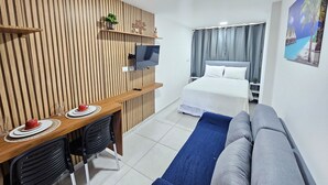 A hotel room with a wooden slat wall, a TV mounted on it, a bed with white linens, a blue sofa, and a small table with a red cup and saucer. - Eco Mirage Residence (Ipojuca)