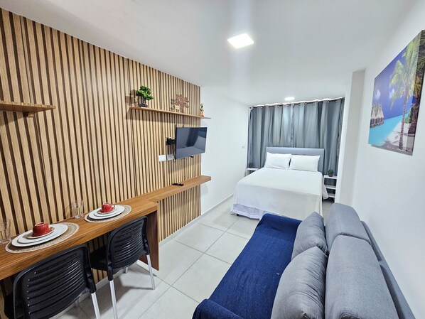 A hotel room with a wooden slat wall, a TV mounted on it, a bed with white linens, a blue sofa, and a small table with a red cup and saucer. - Eco Mirage Residence (Ipojuca)