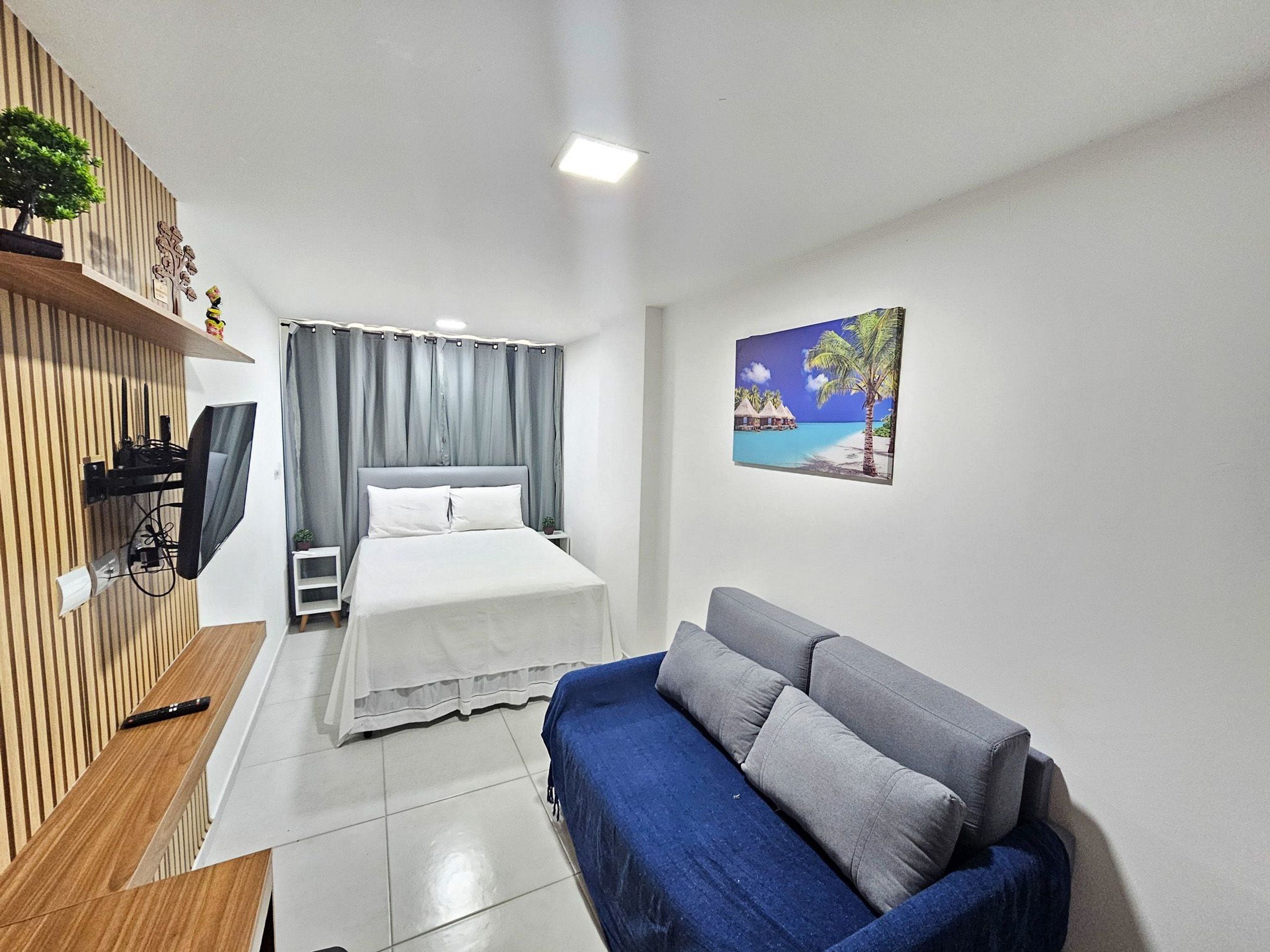 A bedroom with a bed, a TV mounted on the wall, a blue sofa, and a painting of a beach scene.