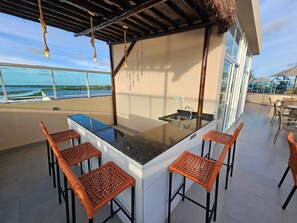 Eine Dachterrasse mit einer Bar, deren Arbeitsfläche aus Glas ist, umgeben von Korbstühlen und mit Blick auf das Meer und ein Sportfeld.