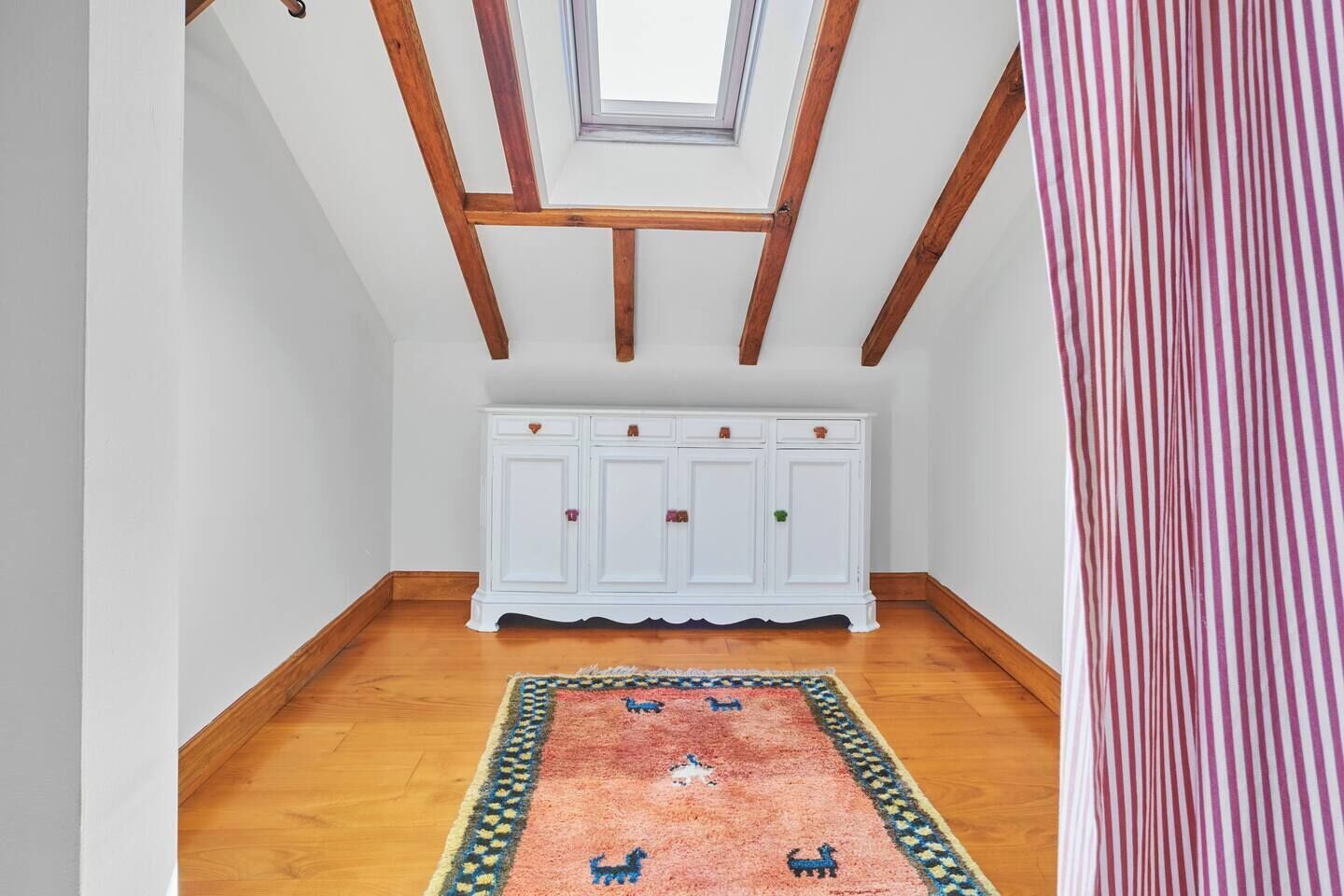 A room with a sloped ceiling, wooden beams, a white cabinet, and a patterned rug.