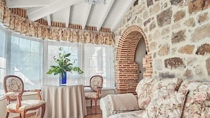Une pièce avec un mur en pierre, une table ronde entourée de chaises, un canapé à motifs floraux et une grande fenêtre avec des rideaux.