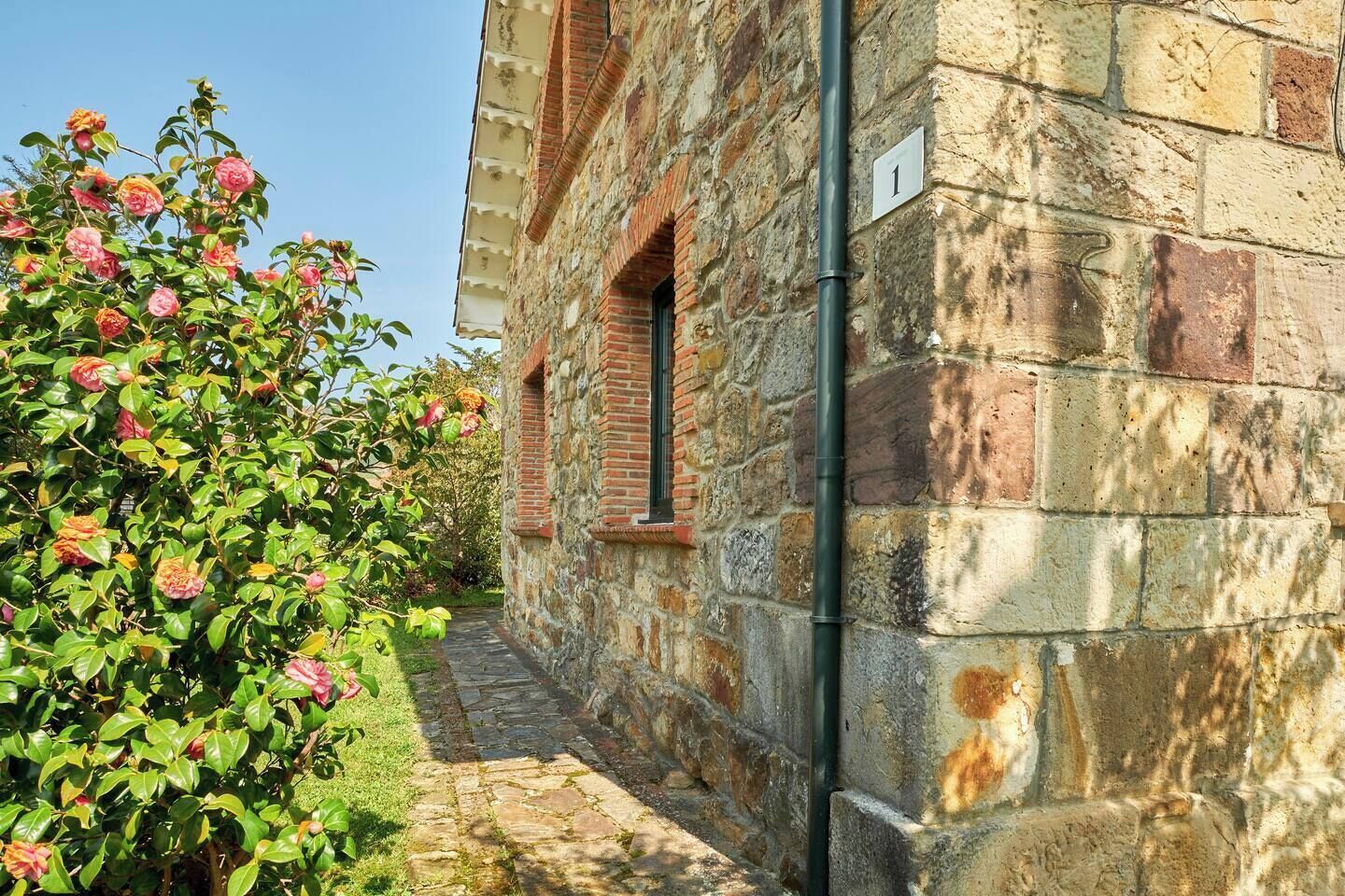 A stone building with a narrow stone pathway leading to a door, surrounded by lush greenery and flowering plants.